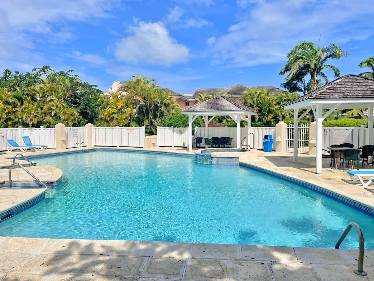 A large swimming pool is surrounded by a spacious deck, featuring comfortable seating areas under shaded pavilions. Lush greenery and palm trees provide a serene backdrop, enhancing the inviting atmosphere. The clear blue water glistens in the sunlight, creating a refreshing outdoor space.