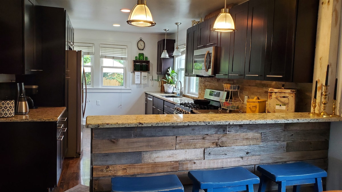 A modern kitchen is equipped with dark cabinetry and granite countertops. Stainless steel appliances are positioned alongside a wooden accent wall. Three blue stools are placed at the kitchen island, complementing the functional design. Natural light enters through nearby windows, enhancing the inviting setting.