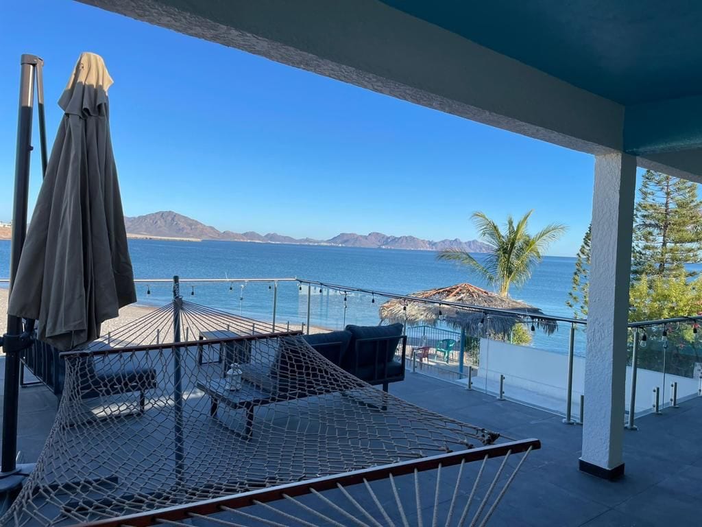 A spacious terrace offers a stunning view of the sea, featuring a hammock and outdoor seating. Clear glass railings provide an unobstructed sightline to the tranquil water and distant mountains, while palm trees and a thatched-roof palapa enhance the natural surroundings.