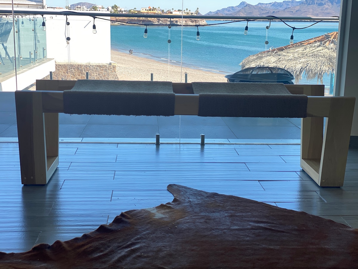 A minimalist wooden bench is positioned by a large glass window, offering a clear view of the beach and ocean. Natural light illuminates the space, and the surface is complemented by a textured fabric runner. Scenic mountains in the distance enhance the overall tranquility.