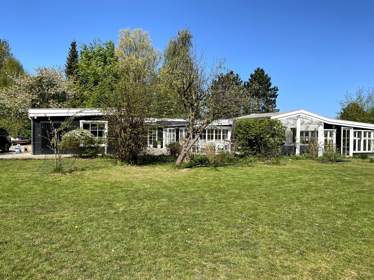 The exterior of the holiday home is displayed, featuring a modern design amid a spacious green lawn. Large windows provide natural light, and lush trees and shrubs enhance the surrounding landscape. The clear blue sky and well-maintained gardens contribute to a serene outdoor setting.