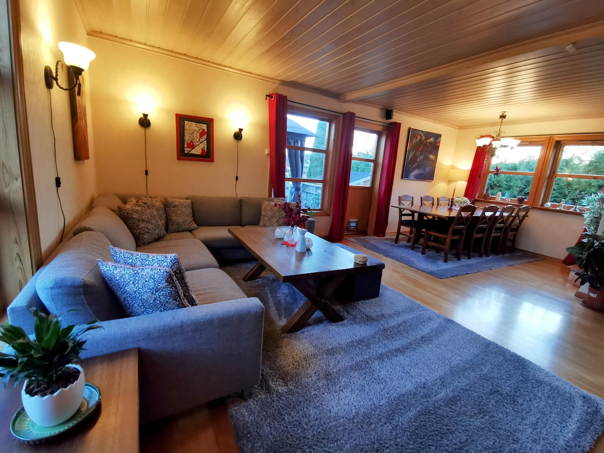 A spacious living area features a comfortable sectional sofa with decorative cushions, complemented by a wooden coffee table. A dining table with multiple chairs is positioned near large windows, providing a view of the outdoors. Soft lighting enhances the inviting ambiance.