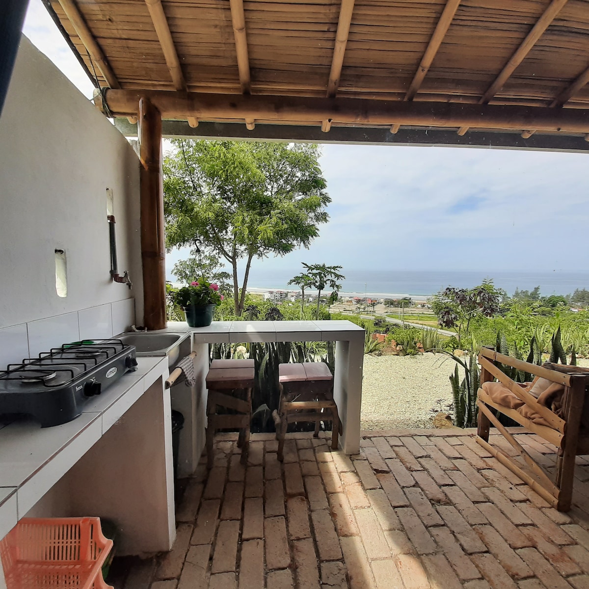An outdoor kitchen area is shown, featuring a simple countertop with a stove and two wooden stools. Lush greenery and a view of the ocean are visible in the background, providing a serene atmosphere for cooking and dining.
