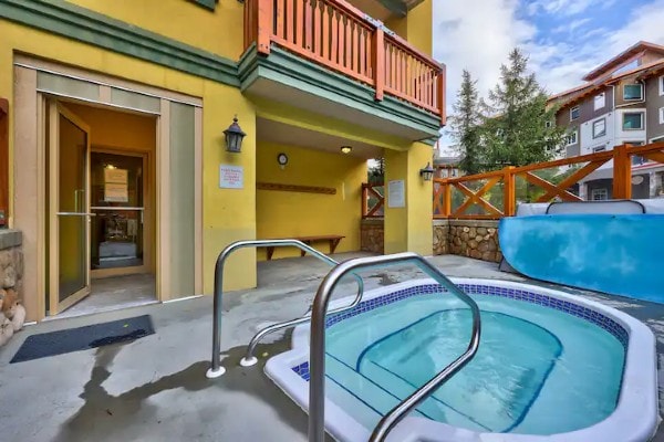 An outdoor hot tub is situated next to a colorful building with a wooden balcony. The entrance to a changing area is visible, featuring a bench and a light fixture. Surrounding trees enhance the serene outdoor setting.