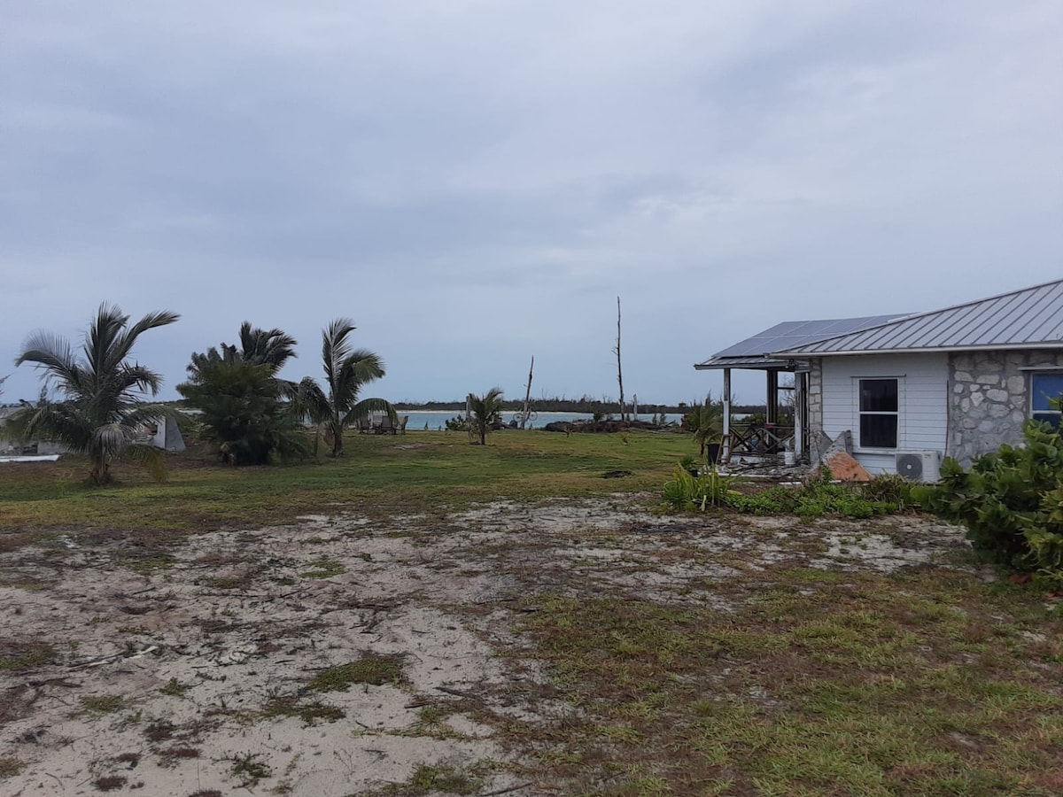 A spacious outdoor area showcases lush greenery and sandy patches, leading to a view of the water in the distance. Palm trees sway gently in the breeze, while a single-story house with a metal roof is positioned to the right, offering a sense of coastal tranquility.