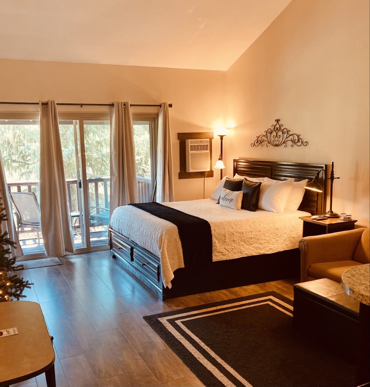 A spacious studio features a king bed with a decorative headboard, complemented by bedside lamps. Natural light filters through large sliding doors that lead to a private deck, framed by curtains. A comfortable loveseat and small dining table add functionality to the cozy interior.