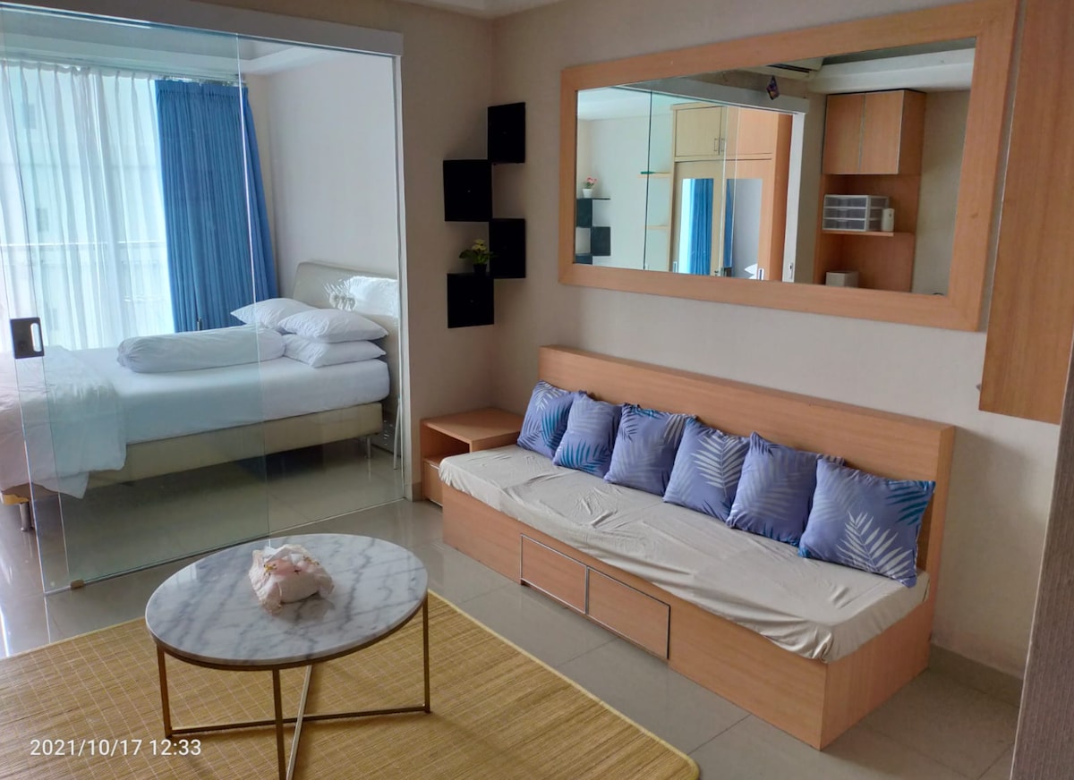 A spacious area is visible featuring a glass partition that separates the living space from the bedroom. A cozy sofa with several blue pillows is positioned against the wall, accompanied by a marble-top coffee table. Light enters through large windows, illuminating the space.