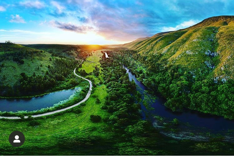 A panoramic view captures lush green valleys and winding rivers beneath an expansive sky. Gentle hills rise in the background, illuminated by the soft glow of the setting sun, reflecting an array of colors across the landscape.