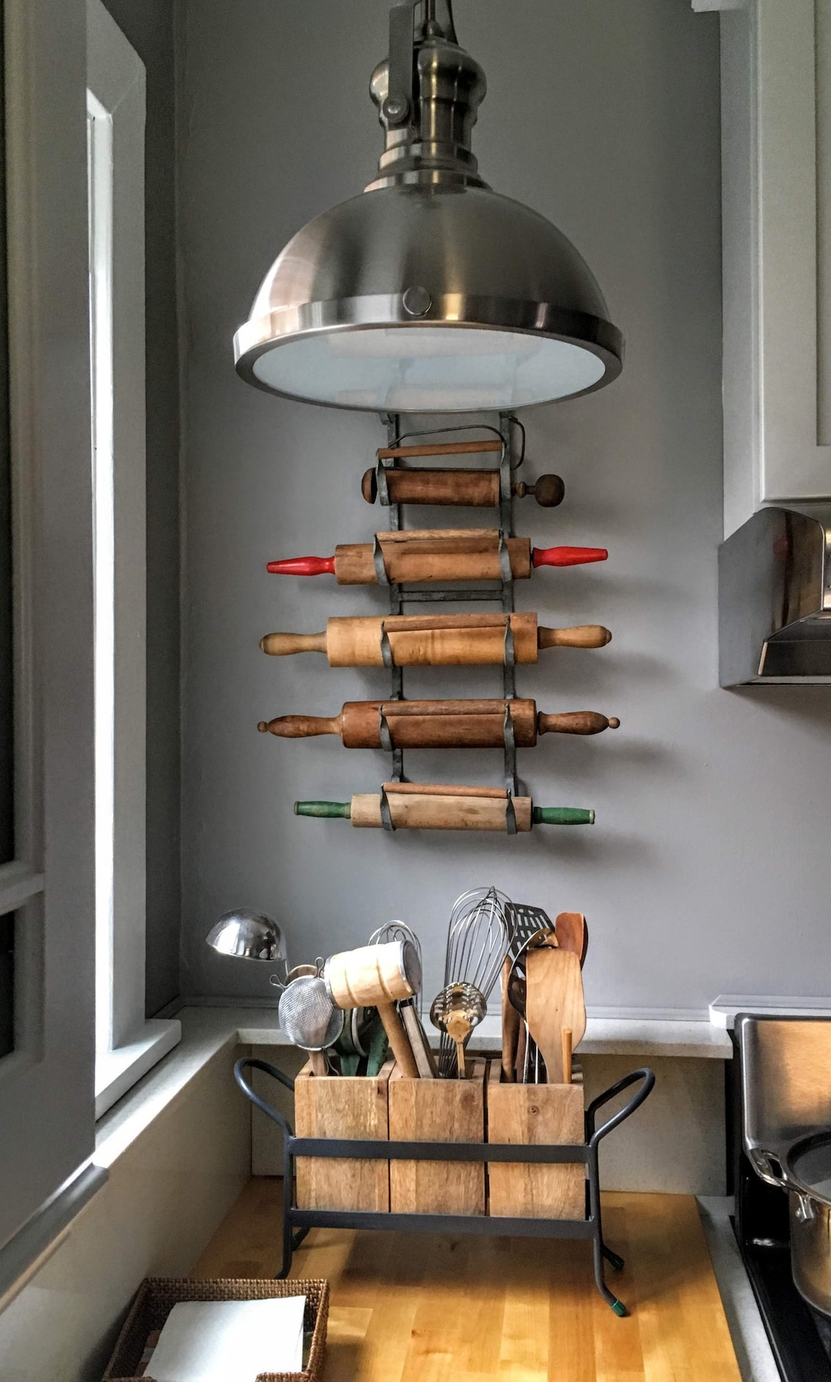A well-organized kitchen corner features a collection of rolling pins hanging on a wall-mounted rack. A metal light fixture provides illumination above. Below, a wooden utensil holder is filled with various kitchen tools, including whisks and wooden spoons, enhancing the cooking space.