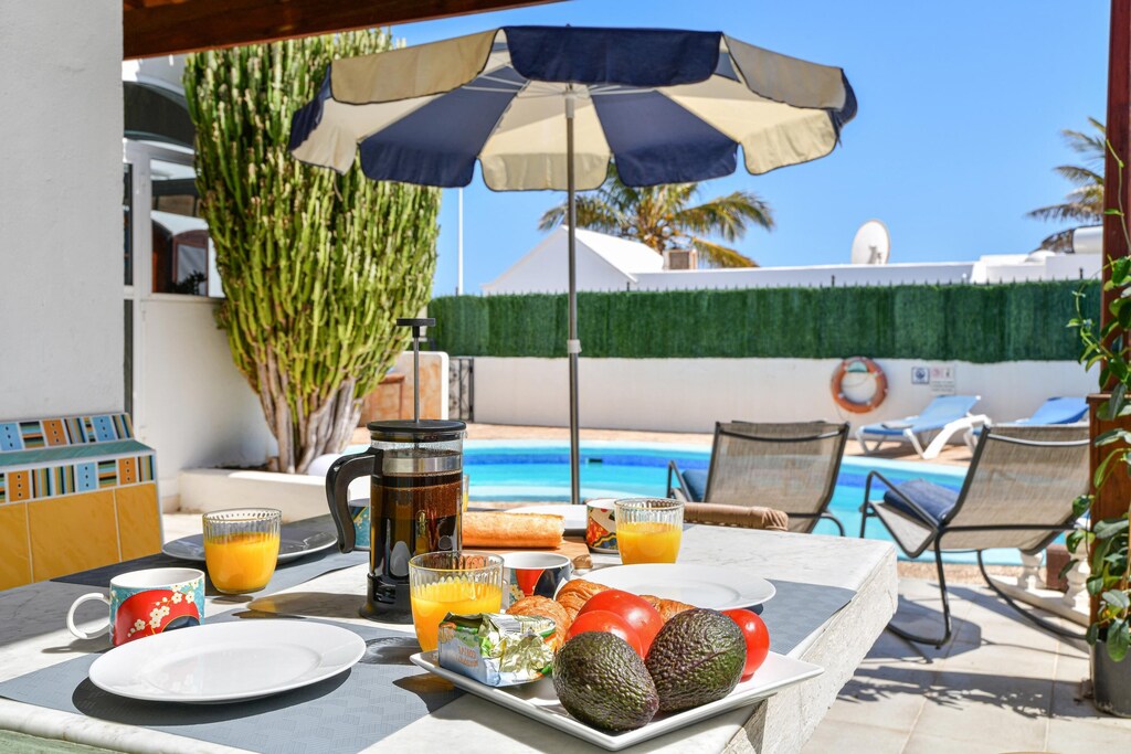 A dining table is set with plates, coffee, and juice, accompanied by fresh avocados and tomatoes. A sunshade provides shade over the table, while a private pool and palm trees are visible in the background, creating a relaxing outdoor atmosphere.