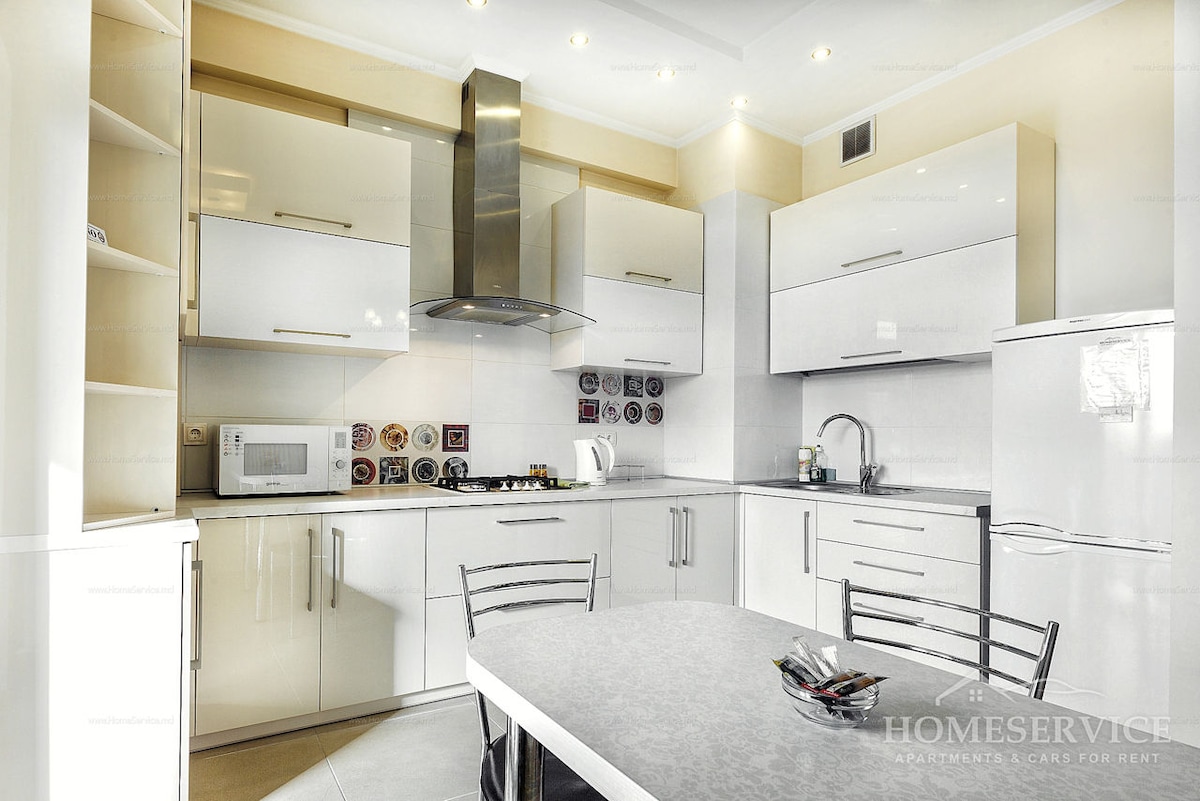 A modern kitchen is equipped with sleek white cabinetry and stainless steel appliances. A round dining table is set at the center, surrounded by four metal chairs. Decorative plates are displayed on the wall, adding a personal touch to the space.