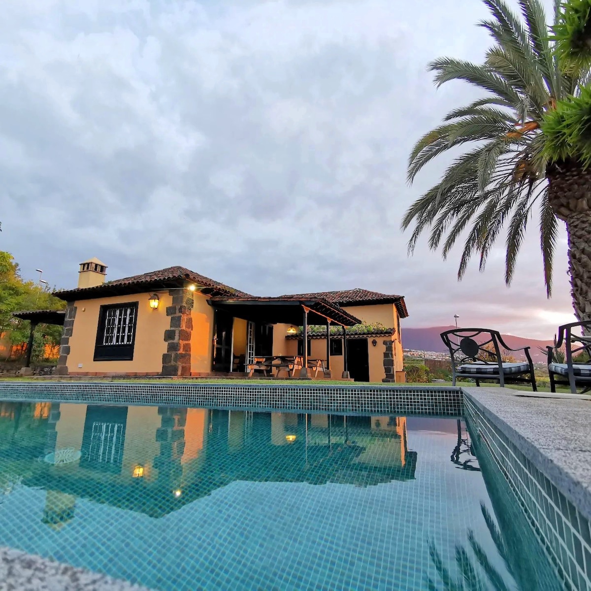 The exterior of a traditionally styled house is reflected in the calm waters of a swimming pool. A palm tree frames the image, while outdoor seating is visible under a covered patio. Soft lighting from the house creates a welcoming ambiance.