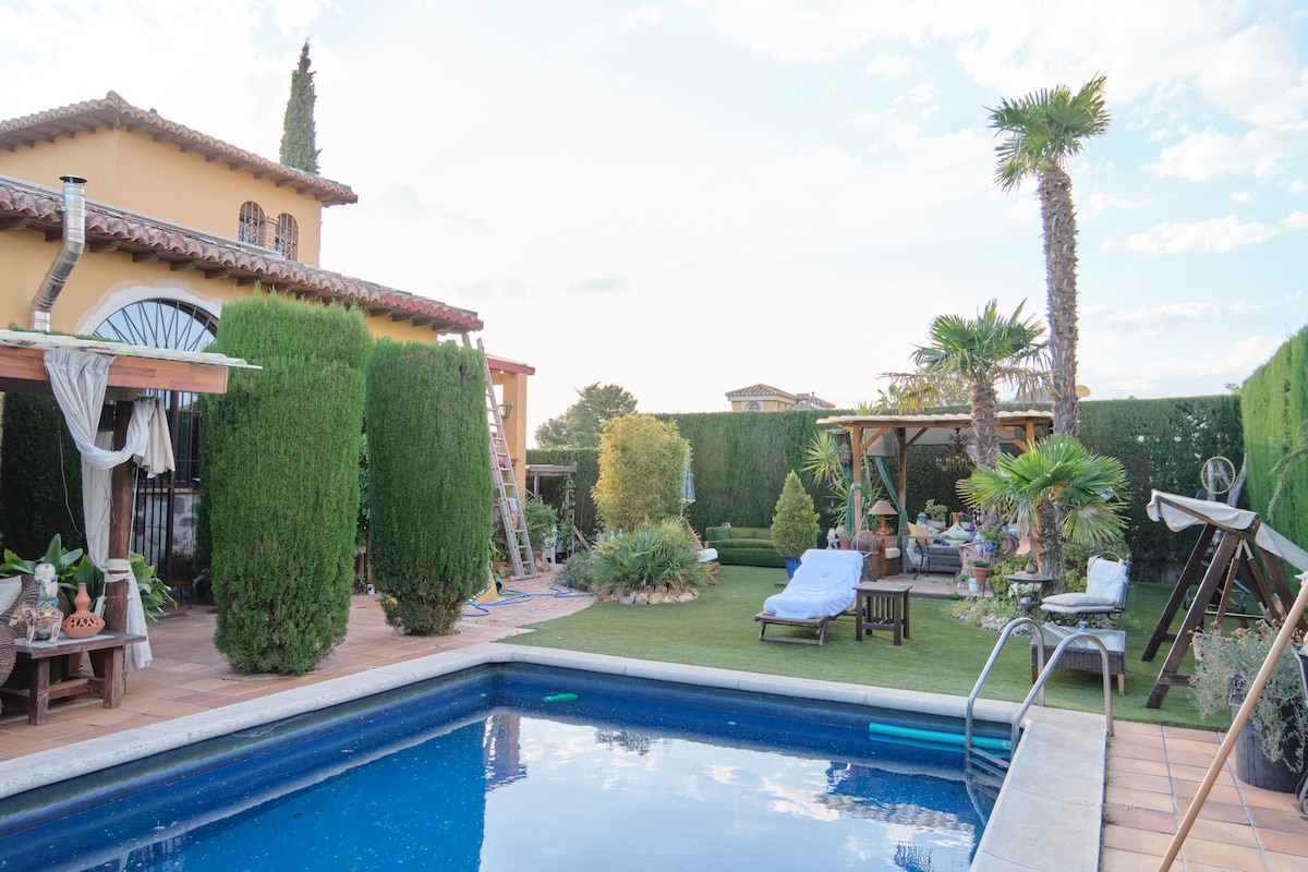 A private swimming pool is surrounded by a lush garden featuring palm trees and various plants. Loungers are positioned near the pool, offering relaxation space. The area includes wooden pergolas and garden art, creating an inviting outdoor setting.