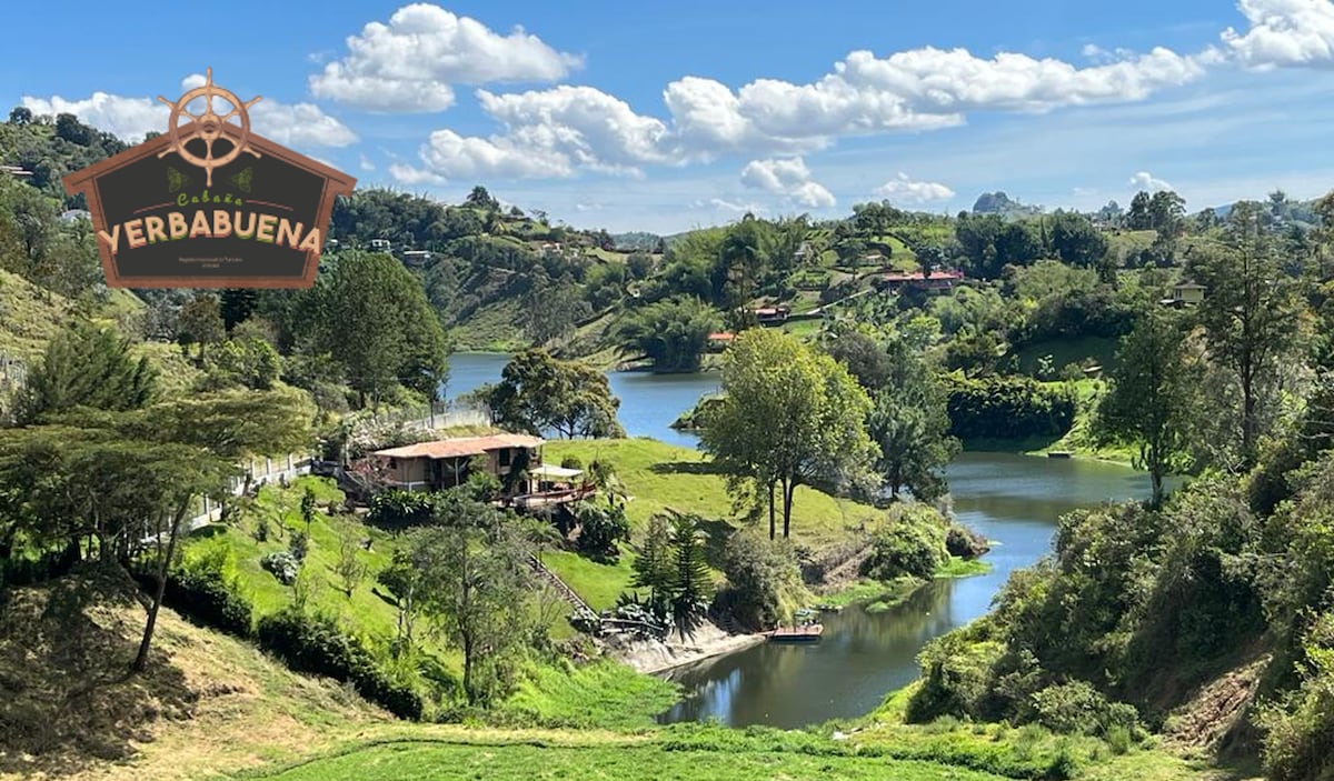 A scenic view showcases the surrounding landscape, featuring lush greenery and a lake. The vibrant blue sky is dotted with fluffy clouds, while a rustic structure and dock are visible near the water's edge, creating a peaceful outdoor setting.