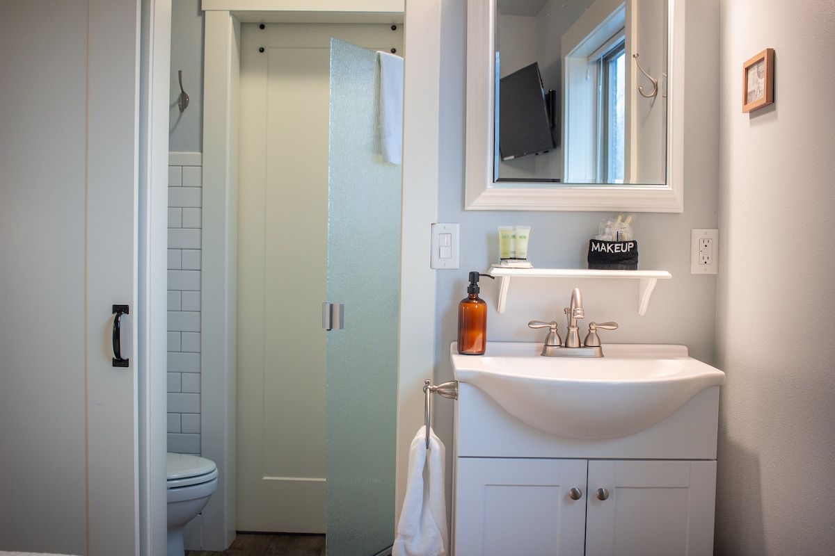 The bathroom features a modern sink with a stylish faucet and an organized shelf with toiletries. A mirror is mounted above the sink, while a frosted glass door provides privacy for the toilet area. Natural light enters through a window nearby.