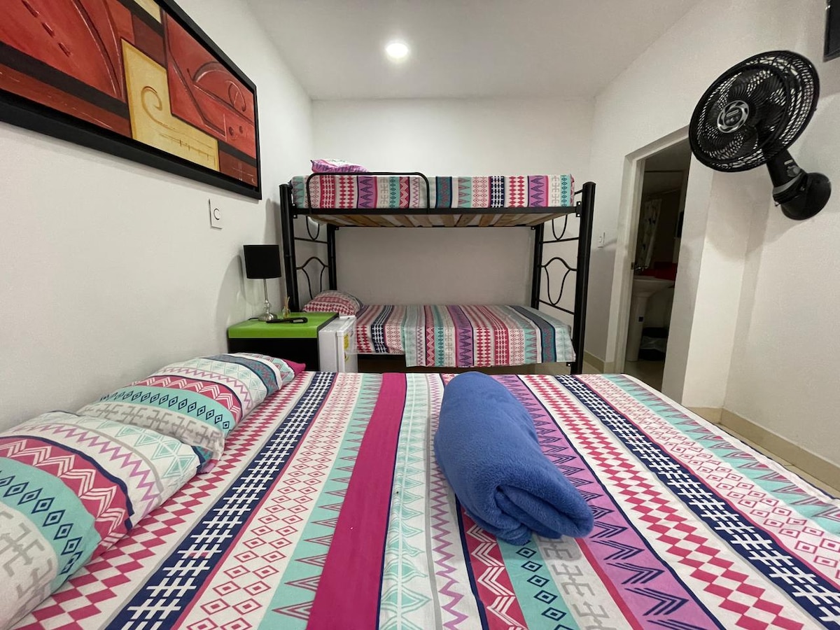 A bedroom features a spacious lower bed with a colorful patterned spread, complemented by a neatly arranged blue towel. Above, a bunk bed is draped in matching patterned bedding. A fan is mounted on the wall, and a small nightstand is positioned beside the lower bed.