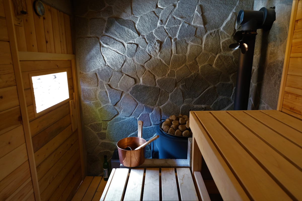 The interior of the sauna is crafted from natural wood, featuring a stone wall that adds texture. A small window allows soft light in, highlighting a wooden bench and a traditional copper bucket beside a blue water container.