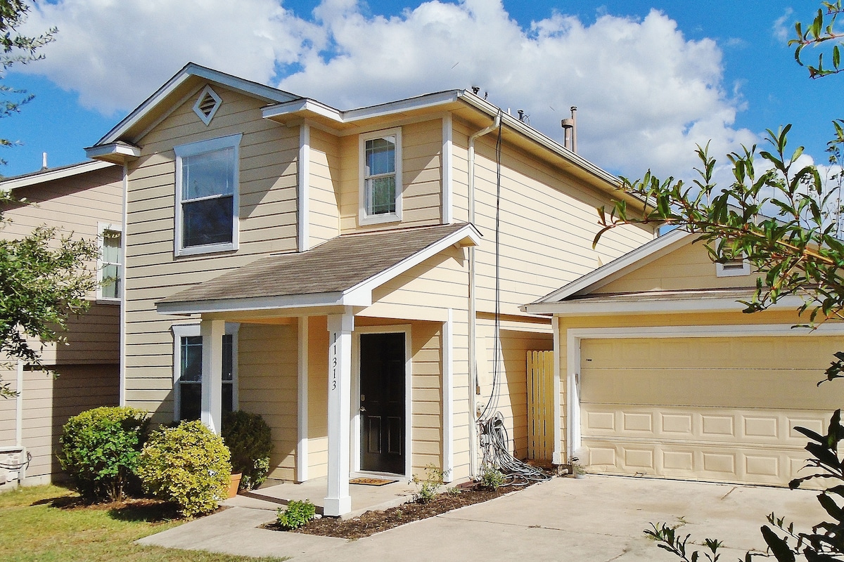 A two-story single-family home is featured, showcasing a light-colored exterior with large windows. A covered front porch offers entry to the home, while a driveway leads to a garage. Lush landscaping adorns the entrance, adding to the welcoming appearance.