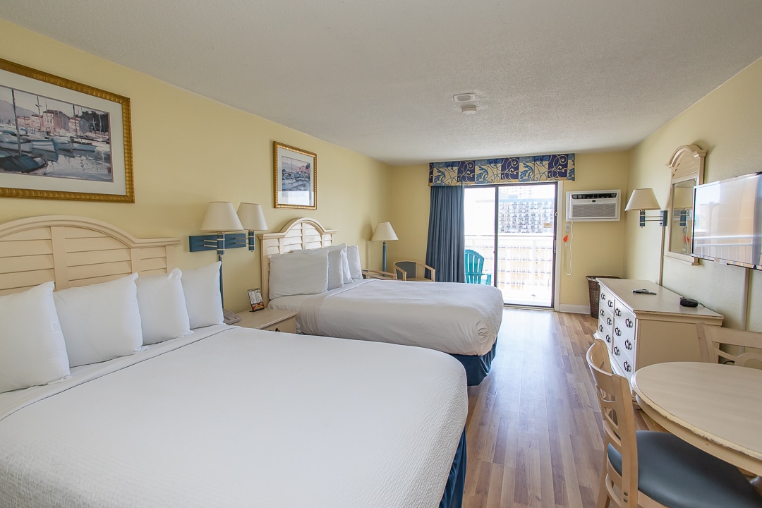 The room features two full beds with crisp white linens, a light wood dresser, and a round table with chairs. A sliding glass door leads to a balcony, allowing natural light to fill the space. A large television is mounted on the wall opposite the beds.