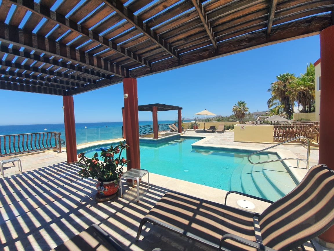 An outdoor infinity pool reflects the sky, surrounded by a spacious deck with lounge chairs. Palapas offer shaded seating, and palm trees add a tropical feel. The expansive ocean view completes this inviting space, enhancing opportunities for relaxation and enjoyment.