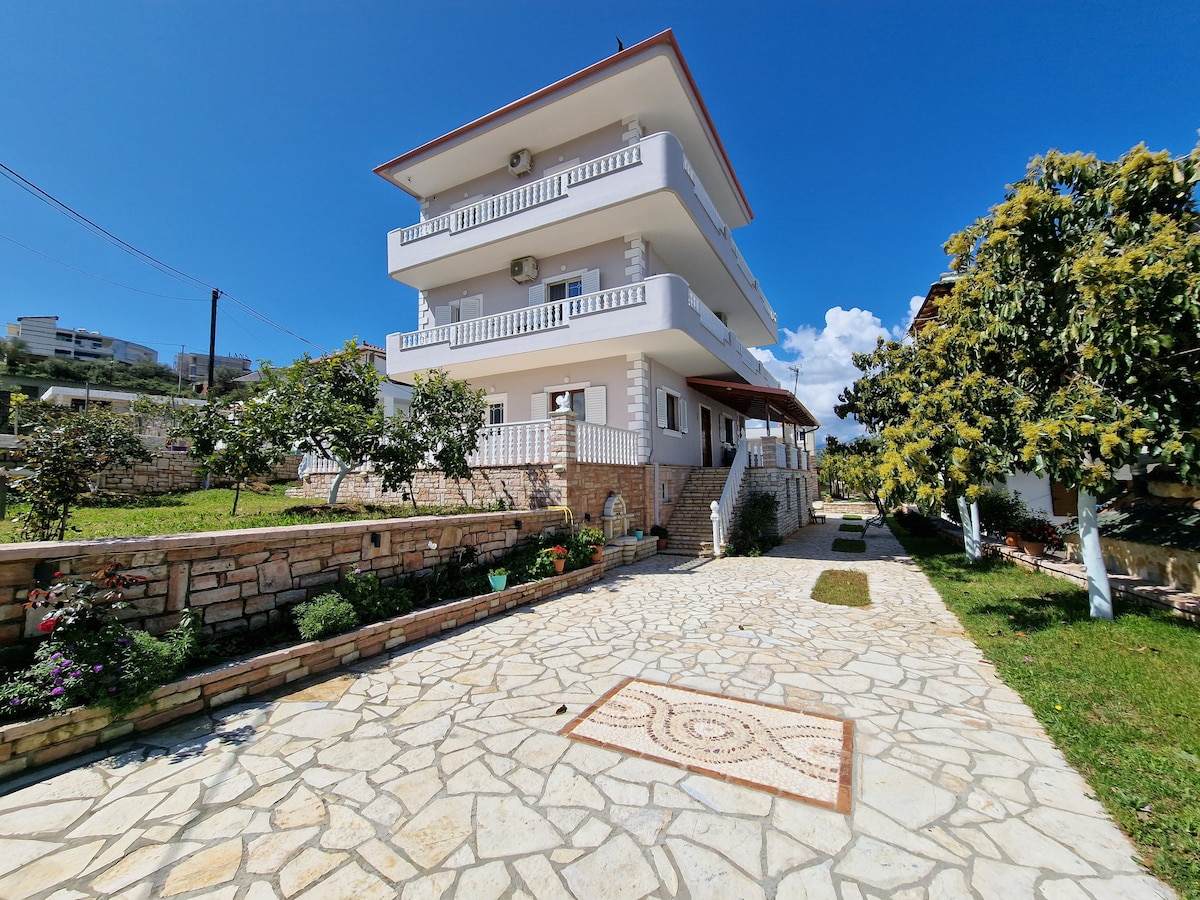 A newly constructed three-story house is shown, featuring a stone pathway leading up to its entrance. Surrounding greenery includes well-maintained gardens and fruit trees. The sky is clear and blue, enhancing the house's bright, welcoming facade.