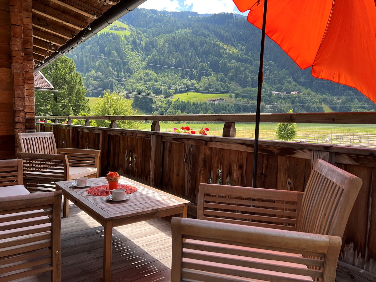 A spacious balcony is shown, furnished with wooden seating and a table. An orange umbrella provides shade, while potted flowers add a colorful touch. The surrounding landscape features lush greenery and distant mountains under a clear blue sky.