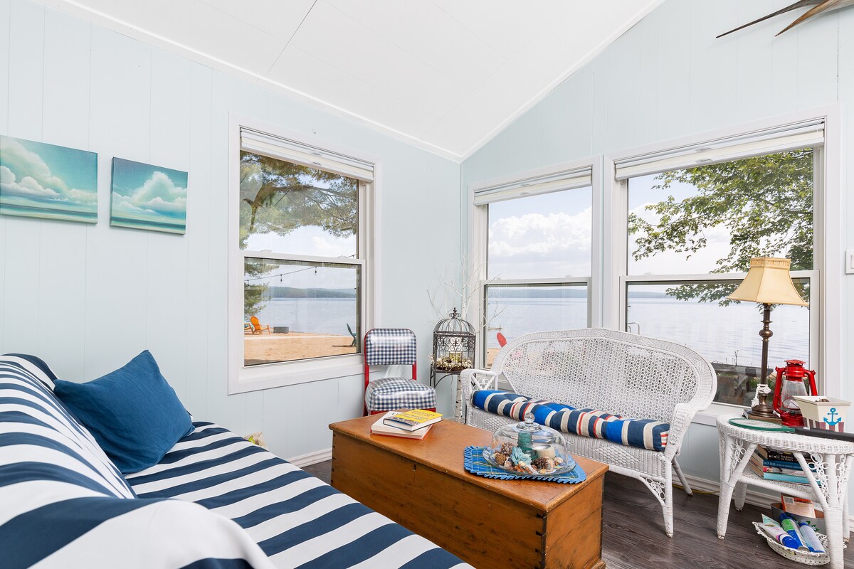 A light-filled sitting area features large windows offering a view of the water. A comfortable sofa with nautical stripes is complemented by woven chairs. A wooden coffee table holds decorative items, and a lamp provides soft lighting, enhancing the space's cozy ambiance.