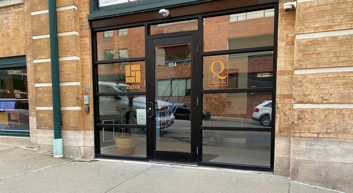 The entrance to the building features large glass doors framed by metal accents, showcasing a clean and modern design. The facade highlights textured brick walls and a potted plant positioned near the doorway, inviting guests into the space.
