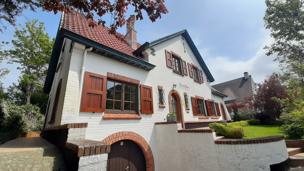 The charming villa features a white brick exterior with wooden shutters. A sloped red roof is complemented by well-maintained landscaping, including trees and shrubs. A staircase leads to the front entrance, which is adorned with decorative elements and a welcoming atmosphere.