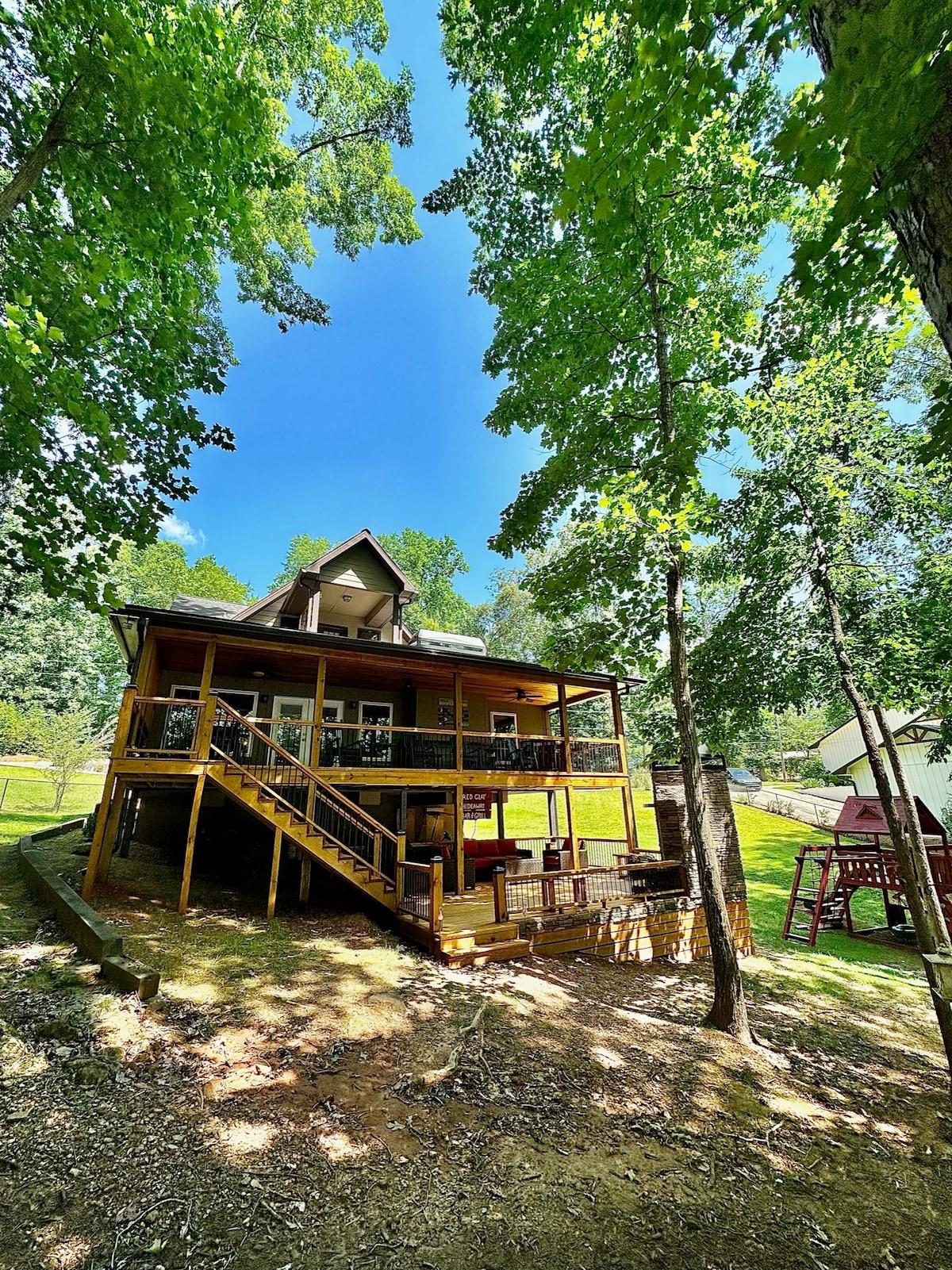 A two-story home with a yellow exterior is surrounded by lush green trees. A spacious deck with railings is visible on the lower level, while steps lead up to the entryway. Bright blue skies are seen above, contrasting with the vibrant greenery.
