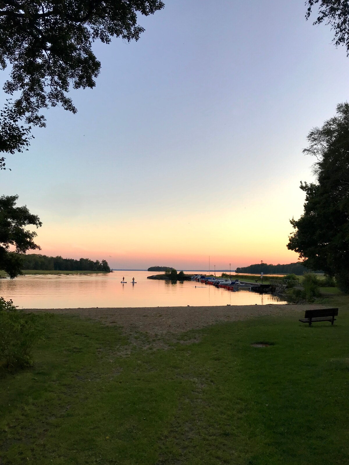 A serene view of Lake Bolmen at sunset captures the tranquil waters reflecting soft pastel hues in the sky. The grassy shoreline leads to a sandy beach, with a couple of silhouettes paddling in the distance. Trees line the edge, providing a natural backdrop.