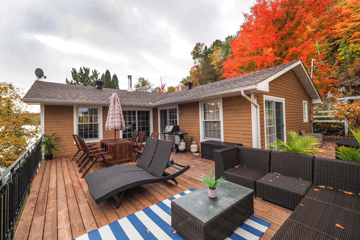 A spacious outdoor deck features a dining table surrounded by wooden chairs, alongside a lounging area with dark, woven furniture. Colorful fall foliage decorates the backdrop, while a large umbrella provides shade. A blue and white striped rug adds a comforting touch to the space.