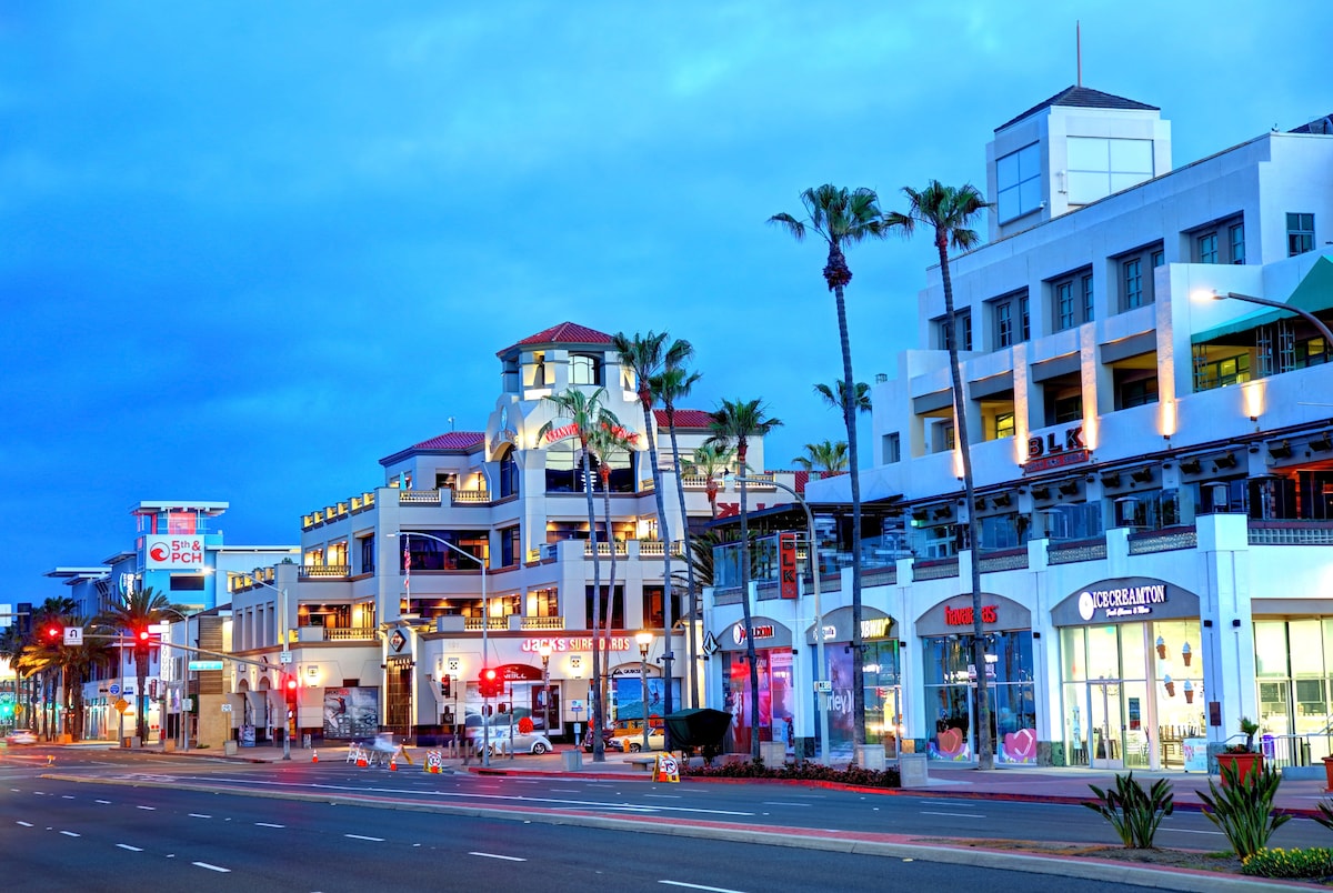 A vibrant street scene in Huntington Beach is captured at dusk, showcasing palm trees lining the roadway and a mix of commercial buildings. Storefronts are illuminated, while multi-level structures with red-tiled roofs create an inviting atmosphere. The sky transitions to a deep blue, enhancing the lively ambiance.