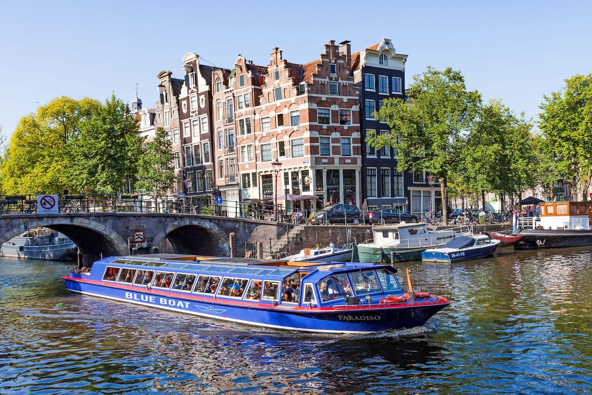 A blue boat glides through the canal, surrounded by tree-lined shores and traditional Dutch architecture on either side. The building facades showcase a mix of colors and styles, while the sunlit water reflects the vibrant scene.