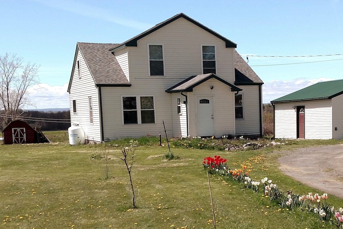 The exterior of a two-story house is shown, featuring a light-colored siding and a symmetrical roof. A welcoming front door is flanked by several windows. A well-maintained yard displays patches of colorful flowers, while a red barn structure and a green shed are visible in the background.