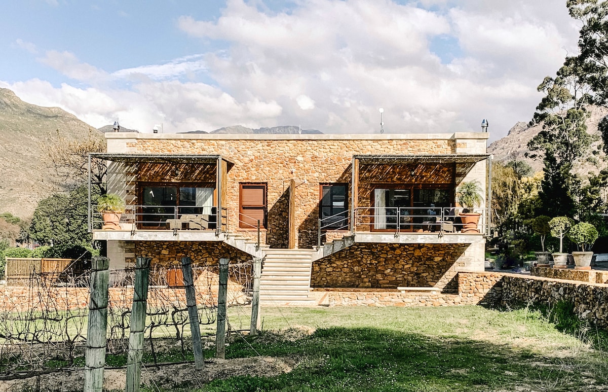 The beautifully structured building features two attached suites with large windows and sliding doors. Each suite is highlighted by an outdoor balcony, providing views of the surrounding landscape. Steps lead to the entrance, complemented by natural stone walls and a terraced garden.