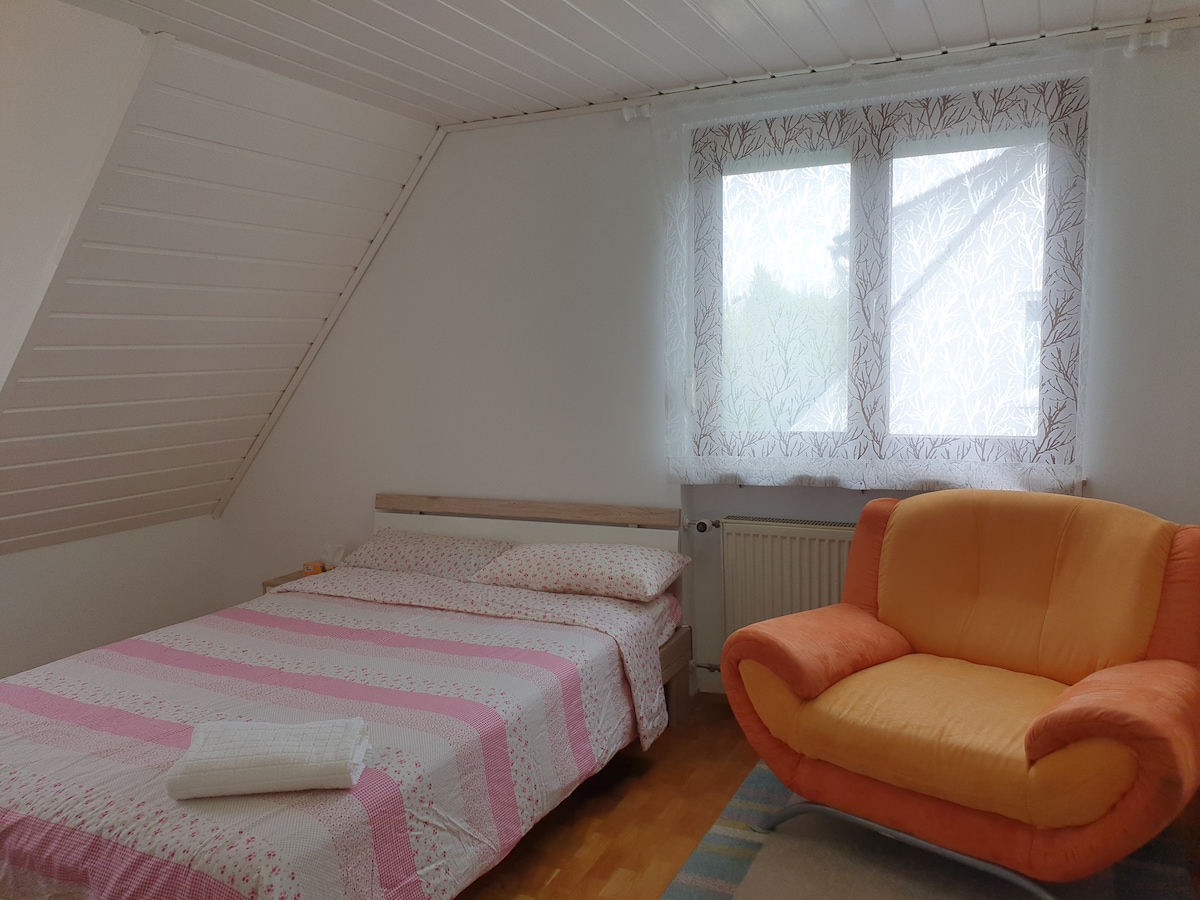 A spacious bedroom is presented, featuring a large bed dressed in a pink and white striped coverlet. An orange sofa adds a pop of color. Natural light filters through two windows adorned with sheer curtains, complementing the wooden flooring and white walls.