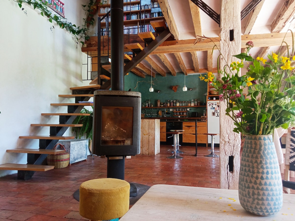 A spacious living area features a wooden staircase leading to an upper level, with a wood-burning stove positioned centrally. The room is adorned with greenery and includes a kitchen area with modern appliances. A vase of fresh flowers adds a pop of color to the table.