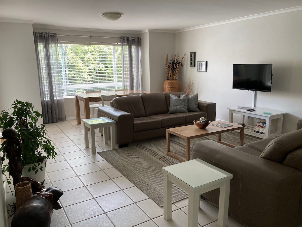 The living area features two large leather couches facing a wall-mounted TV. A coffee table rests on a light area rug, surrounded by light-colored side tables. Natural light enters through large windows, offering a view of greenery outside.