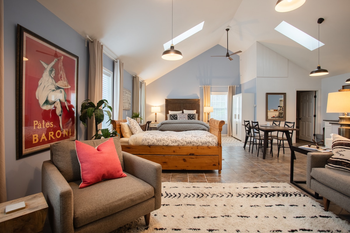 The spacious loft is highlighted by 16-foot ceilings and multiple skylights. A queen bed is positioned near a cozy seating area, complemented by a wooden dining table. Eye-catching artwork adorns the walls, and natural light fills the room through large windows.
