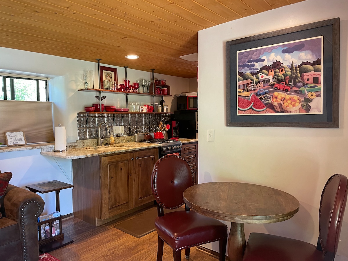 A cozy kitchen area features wooden cabinets and a stone countertop, complemented by red accents. A small dining table is set next to a comfortable seating space, with a colorful artwork hanging on the wall, capturing a vibrant local scene.