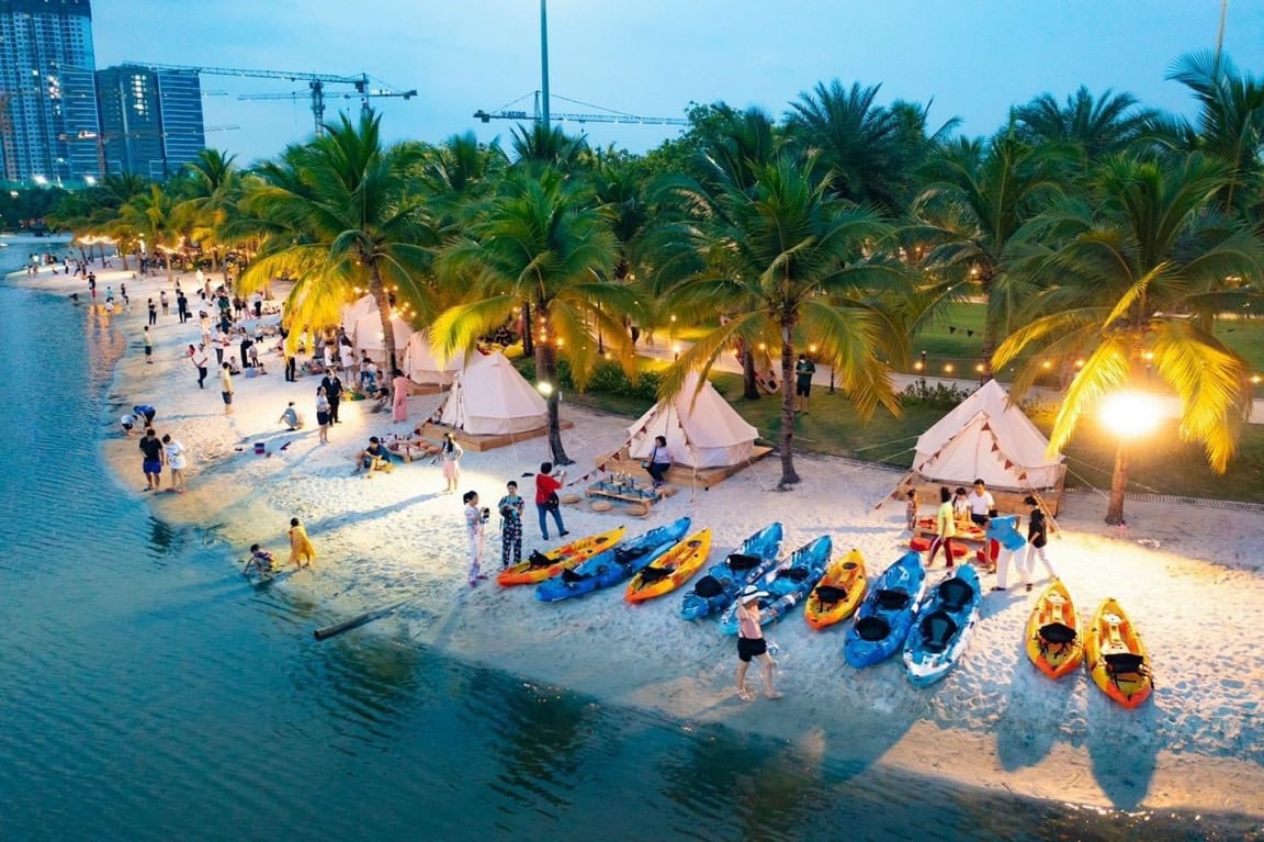 A vibrant beach area showcases an array of colorful kayaks lined along the shoreline. Palm trees provide a lush backdrop, while illuminated tents create cozy spaces for gatherings. People are seen enjoying various activities in the warm evening light by the water's edge.