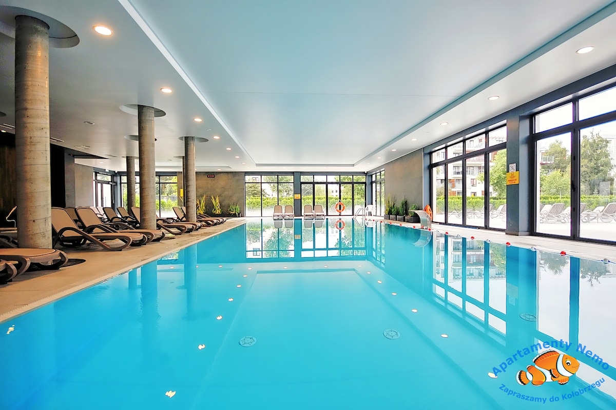 A well-lit indoor swimming pool is surrounded by large windows that provide views of the outside greenery. Comfortable lounge chairs line one side of the pool, enhancing the tranquil atmosphere. The spacious pool area is designed for relaxation and leisure.