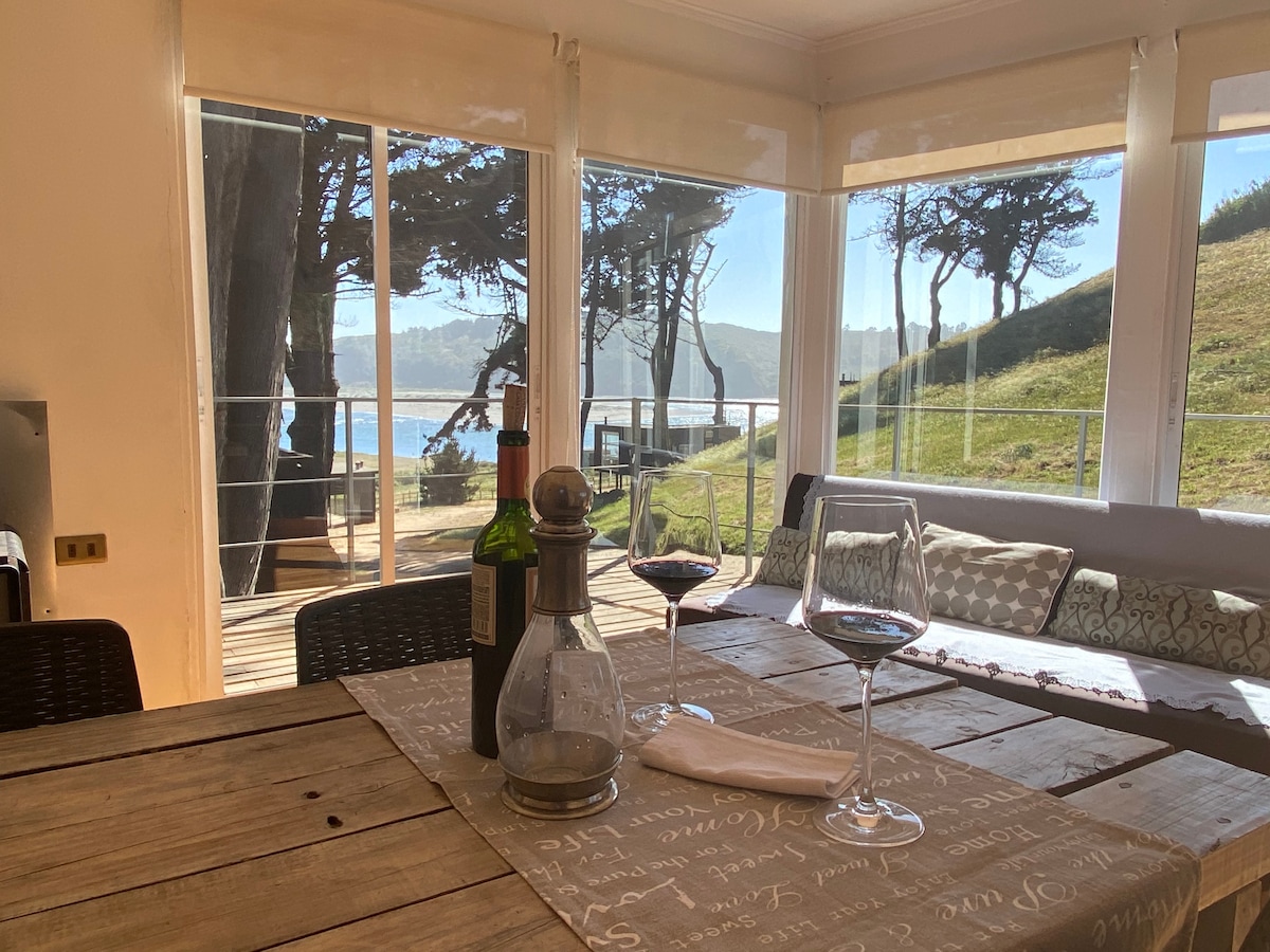 A dining area with a rustic wooden table is set with two wine glasses and a bottle. Large glass doors provide a clear view of the outdoor terrace and lush greenery, enhancing the connection between indoor and outdoor spaces.