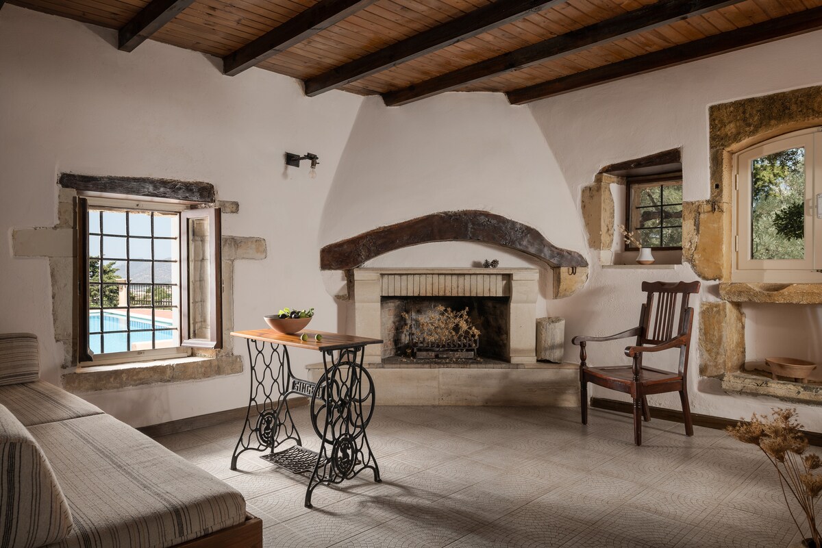 A spacious living area features a central fireplace with a rustic wooden beam above it. Natural light pours in through two windows, one of which offers a view of the swimming pool. A simple wooden chair and a small table enhance the inviting ambiance.