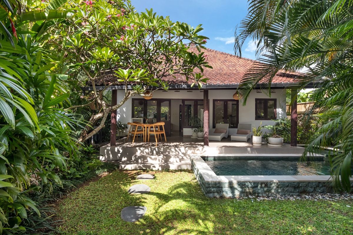 A traditional villa exterior is surrounded by lush greenery, featuring a private pool adjacent to a shaded terrace. The outdoor seating area includes a dining table and lounge chairs, offering a tranquil space to relax. Sunlight filters through the foliage, enhancing the serene environment.