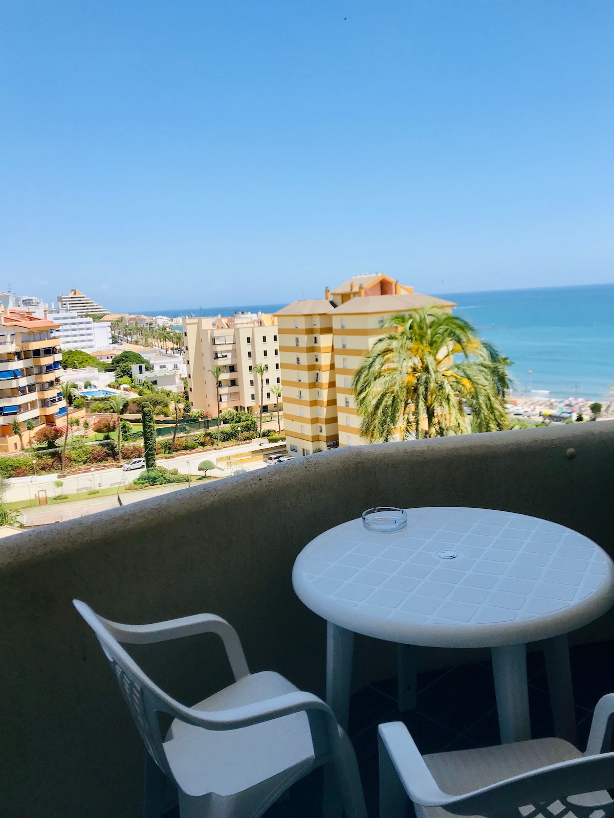 A balcony view features a round white table with two chairs, overlooking the coastline and the ocean. Surrounding buildings and palm trees are visible, enhancing the scene with a sense of place. The sky is clear, providing a bright and open atmosphere.