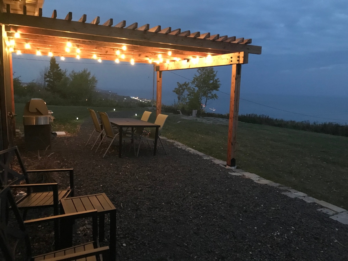 An outdoor patio area is illuminated by string lights, providing a cozy ambiance. A large dining table with chairs is arranged under a wooden pergola, while a grill is positioned nearby. The lawn stretches out beyond, with views of Lake Superior visible in the distance.