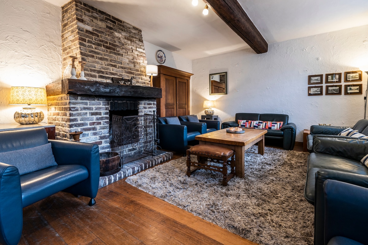 A cozy living area features a rustic stone fireplace, surrounded by comfortable seating arrangements. A central wooden table rests on a soft area rug, while warm lighting from lamps creates an inviting environment. Wall art and framed photographs add visual interest to the space.