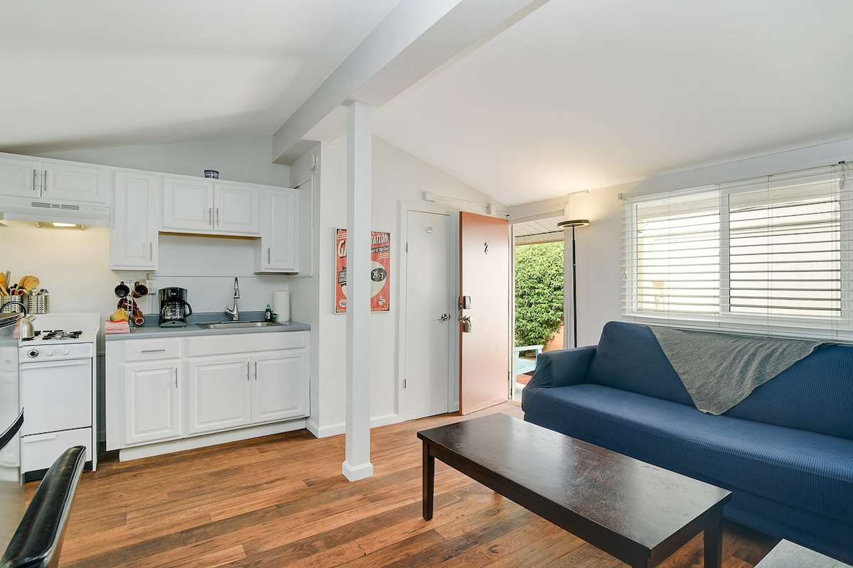 A bright studio space is seen with a kitchen area featuring white cabinetry and modern appliances. A blue couch sits in front of a simple wooden coffee table, while sunlight enters through the window, illuminating the inviting atmosphere and welcoming entryway.